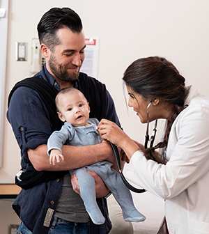 Man holding baby in exam room while healthcare provider listens to baby's chest with stethoscope.