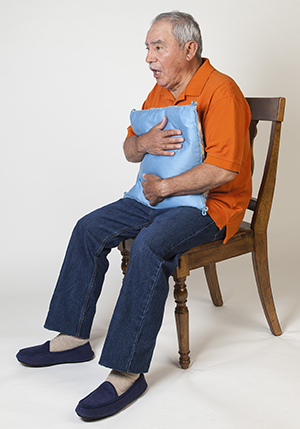Man sitting on chair, holding pillow to chest, coughing.