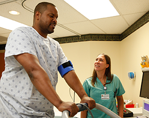 Healthcare provider performing cardiac stress test on man standing on treadmill in exam room.