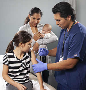 Healthcare provider giving girl injection in arm while woman with baby looks on.