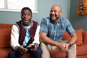 Teen boy with video game controller sitting on couch with man.