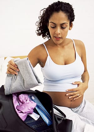 Pregnant person sitting on bed putting items in overnight bag.