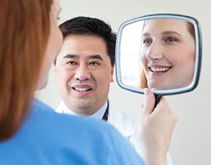 Woman looking in hand mirror with doctor looking on.